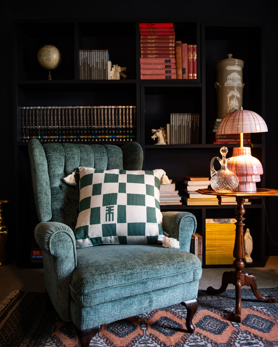 Green armchair with a checkered pillow in a room with bookshelves and a lamp.