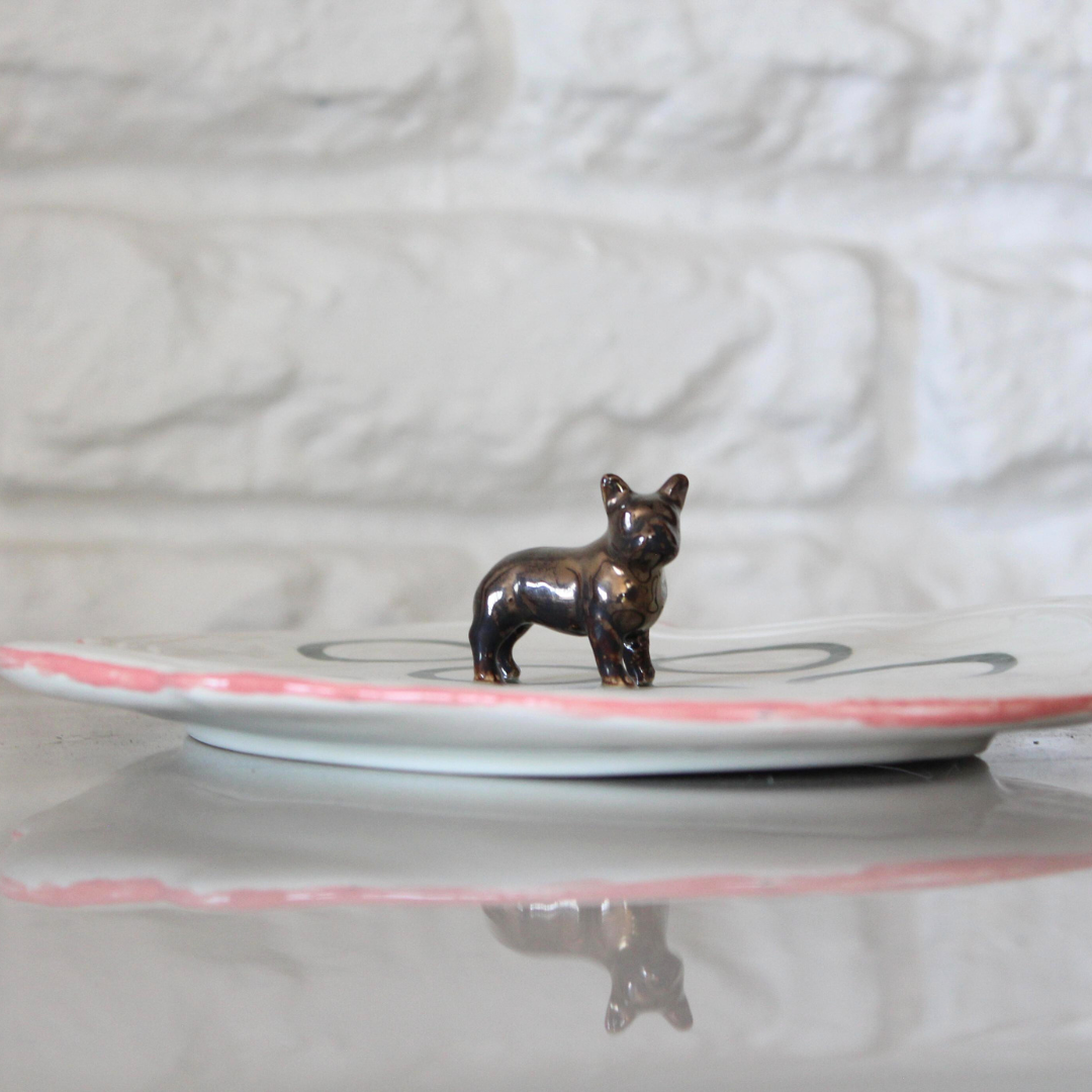 Bronze dog figurine on a white plate with a pink rim against a light gray background