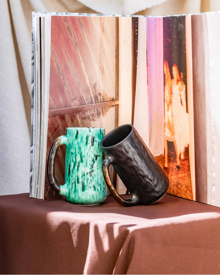 Open book with handmade ceramic mugs on a table with a curtain and outdoor view in the background