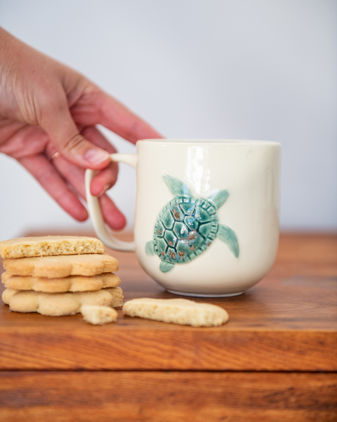 Mug with turtle design held by a hand on a wooden surface with cookies.