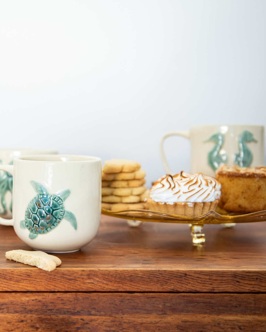 Two ceramic mugs with turtle designs on a wooden surface with cookies and a tart.