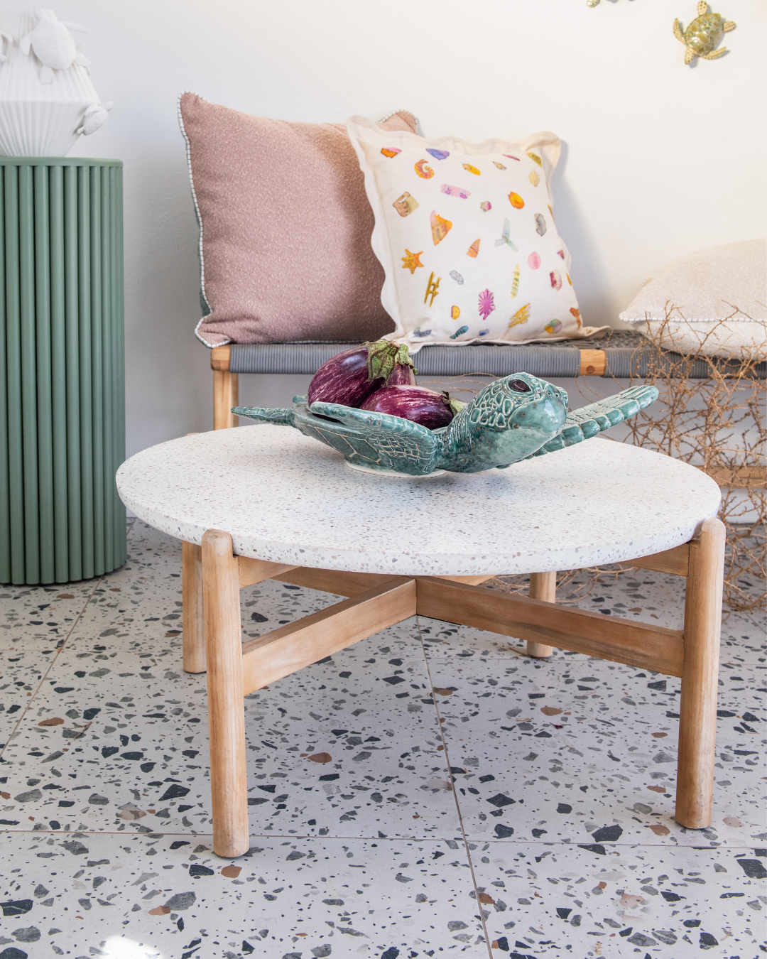 Round coffee table with a large ceramic sea turtle fruit bowl on a textured surface and wooden legs in a room with a radiator and decorative pillows.