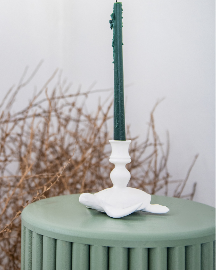 Decorative matt white sea turtle candle stick holders including a green candlestick, on an orange cushion against a white wall.