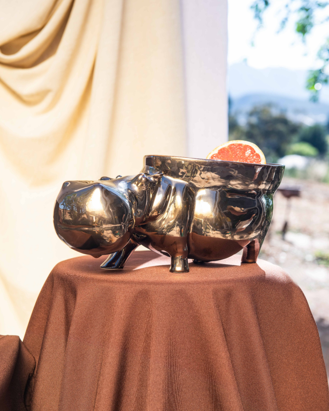Decorative setup with ceramic hippo's with bowls attached to them turning them into fruit bowls on brown tablecloths, a white vase, and fruit bowl against a neutral curtain.