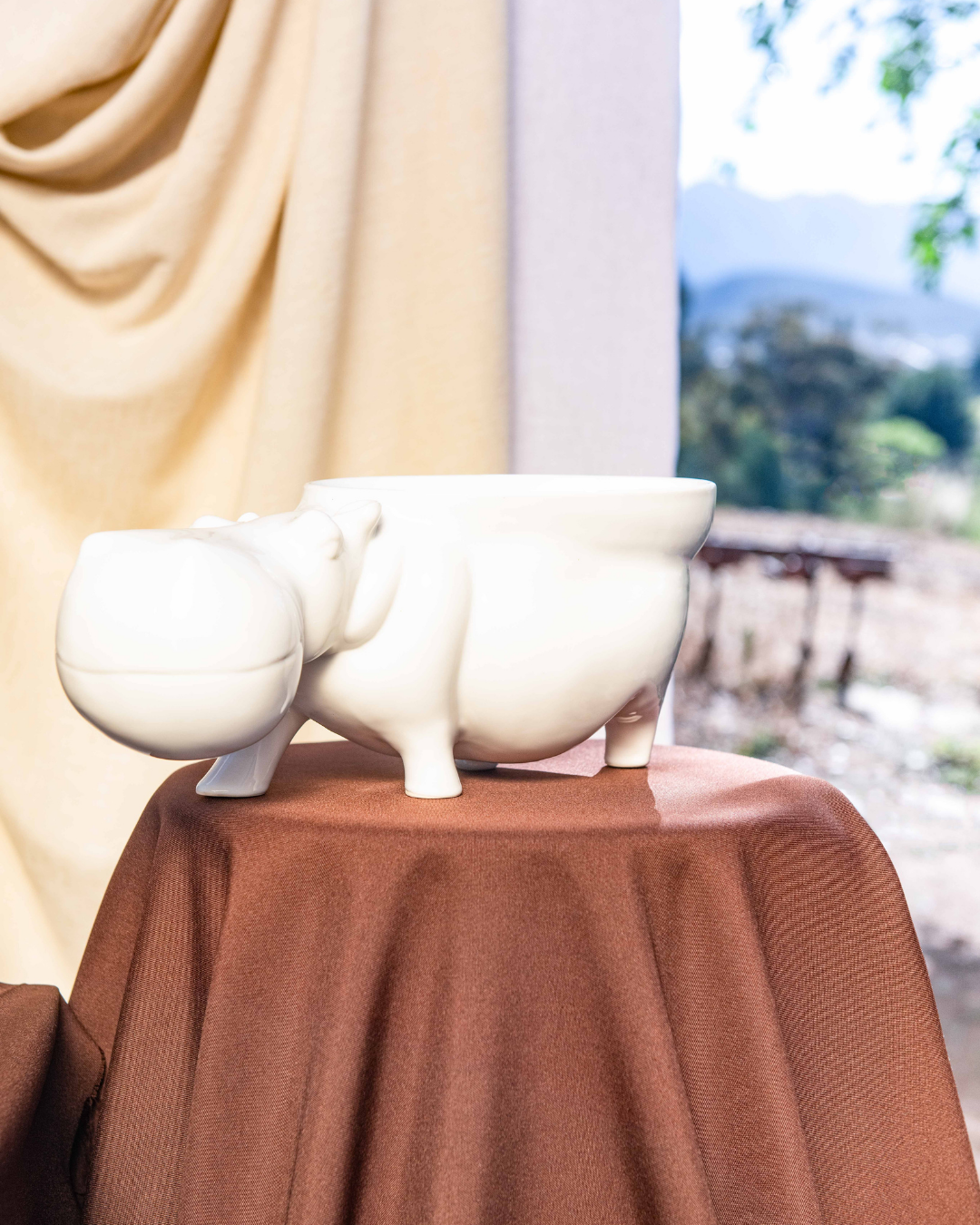 Decorative setup with ceramic hippo's with bowls attached to them turning them into fruit bowls on brown tablecloths, a white vase, and fruit bowl against a neutral curtain.