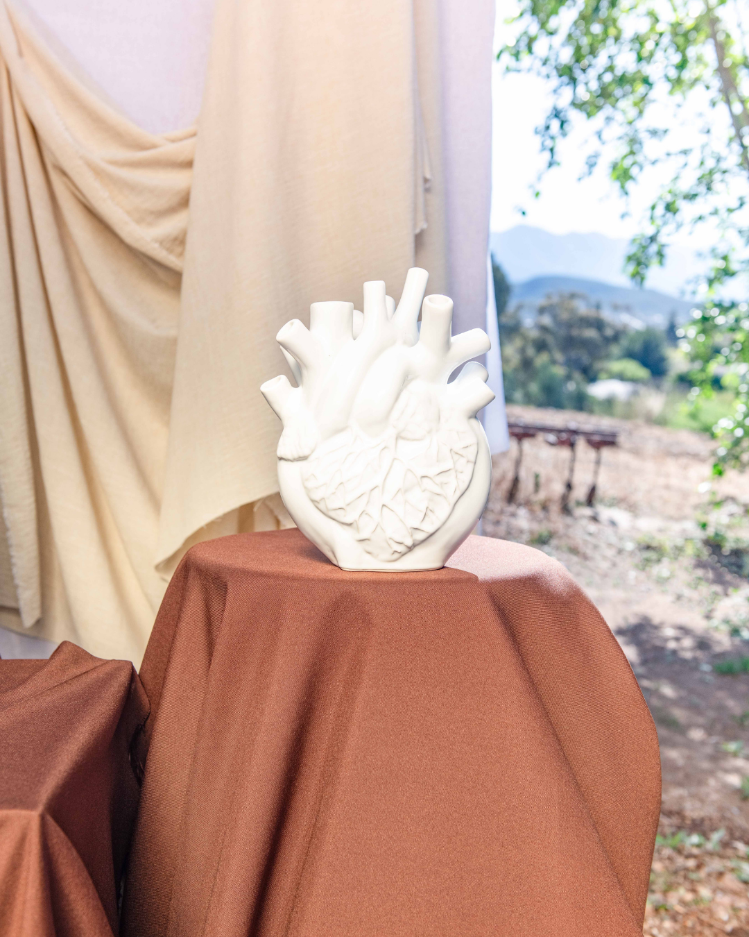 White ceramic heart-shaped vase on a brown surface with beige curtains and outdoor scenery in the background