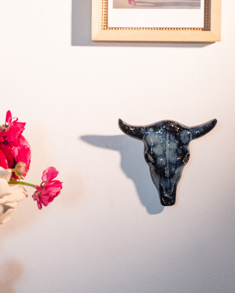 Decorative bull head on a wall with framed pictures above and flowers below.