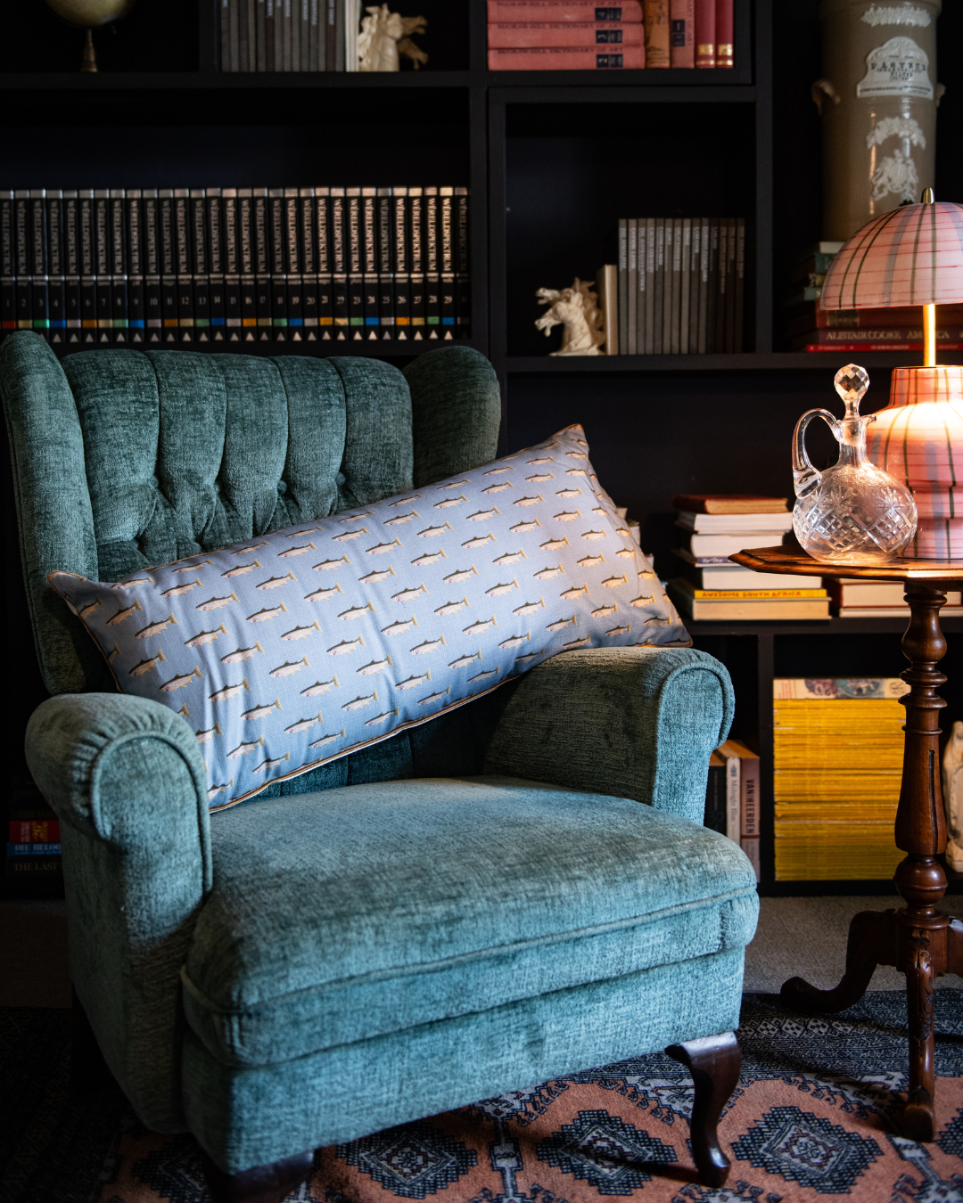 Teal armchair with a decorative pillow in the rainbow trout monogram in faded blue in a cozy room with bookshelves and a table.
