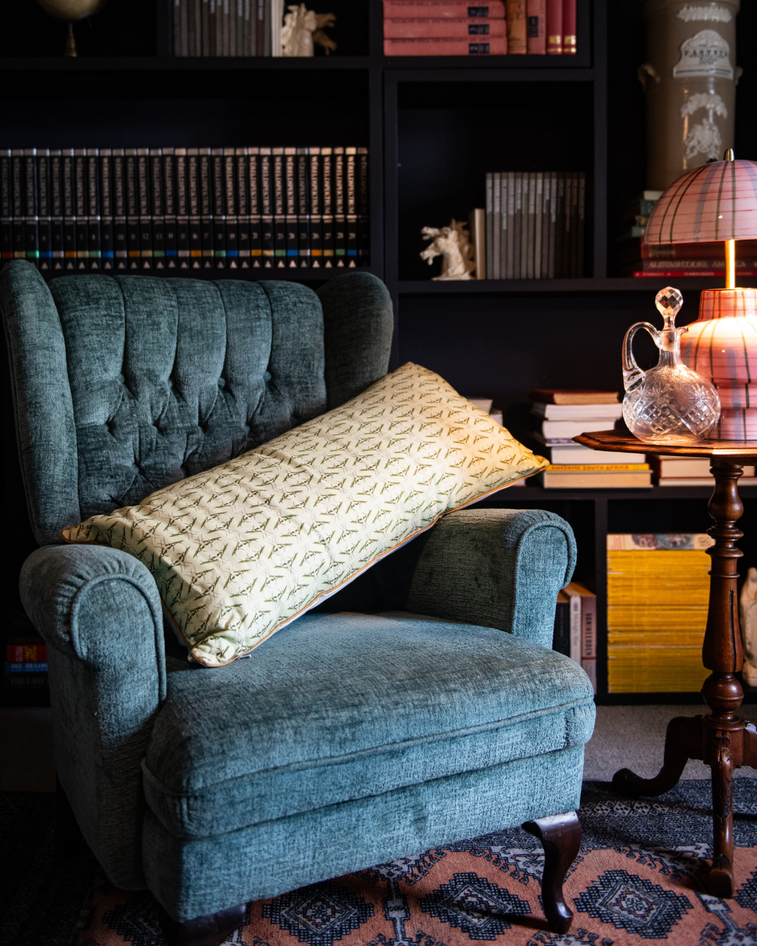 Blue armchair with a patterned pillow in a praying Mantis monogram with a green base in a room with bookshelves and a table.