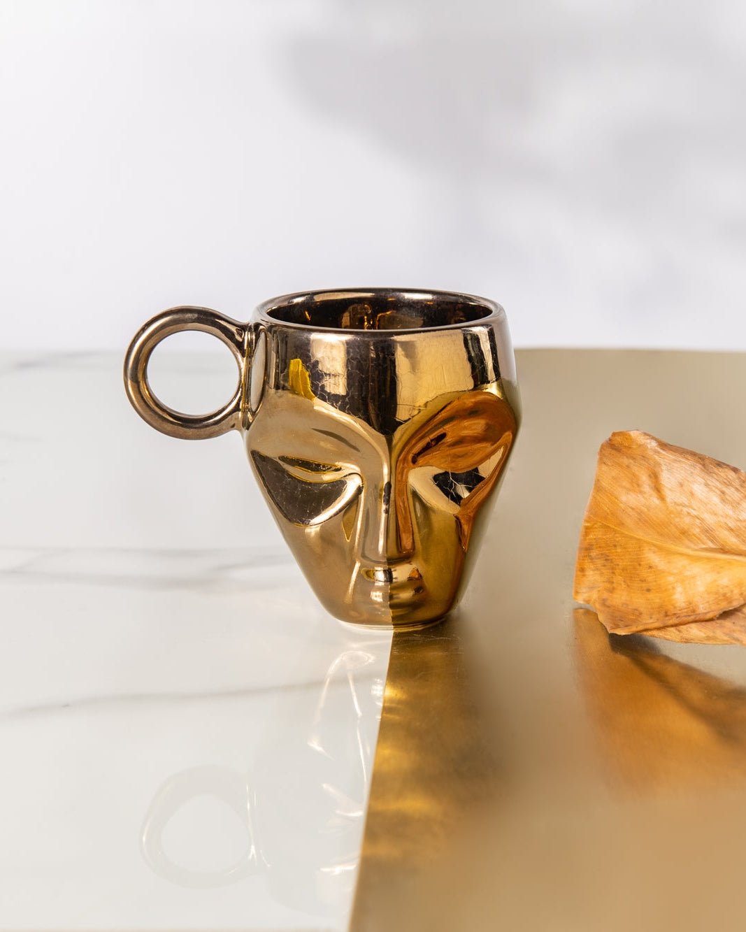Gold mask-shaped mug and decorative leaf on a reflective surface with a white background