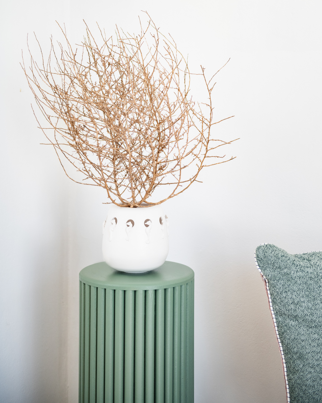 Matt White vase with seahorses with dried branches on a green cylindrical table against a white wall.