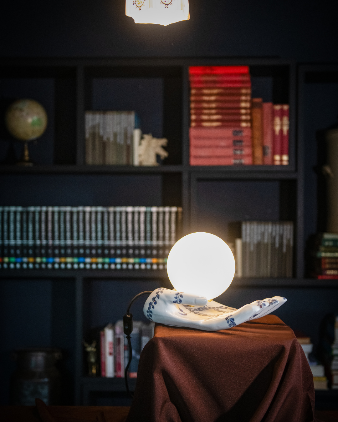 Light bulb exposed on a ceramic light in the shape of a hand with hand painted delf like leaves on the gloss white surface on a table with a bookshelf in the background