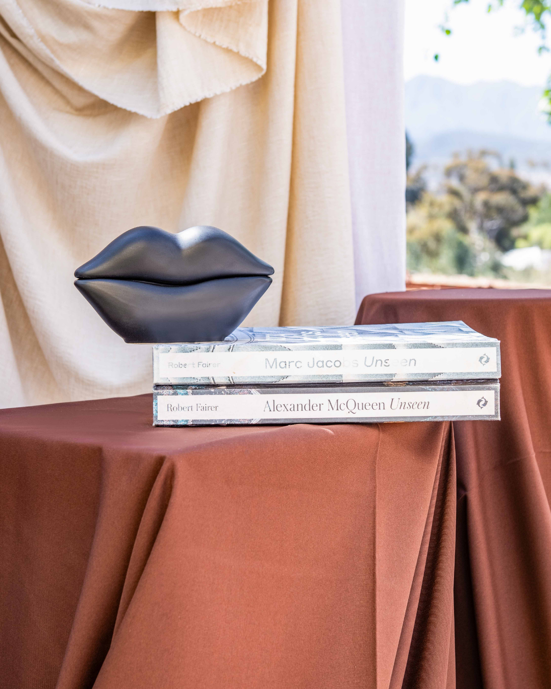 Three books stacked on a table with a metallic lip-shaped object above them, against a beige curtain and outdoor background.