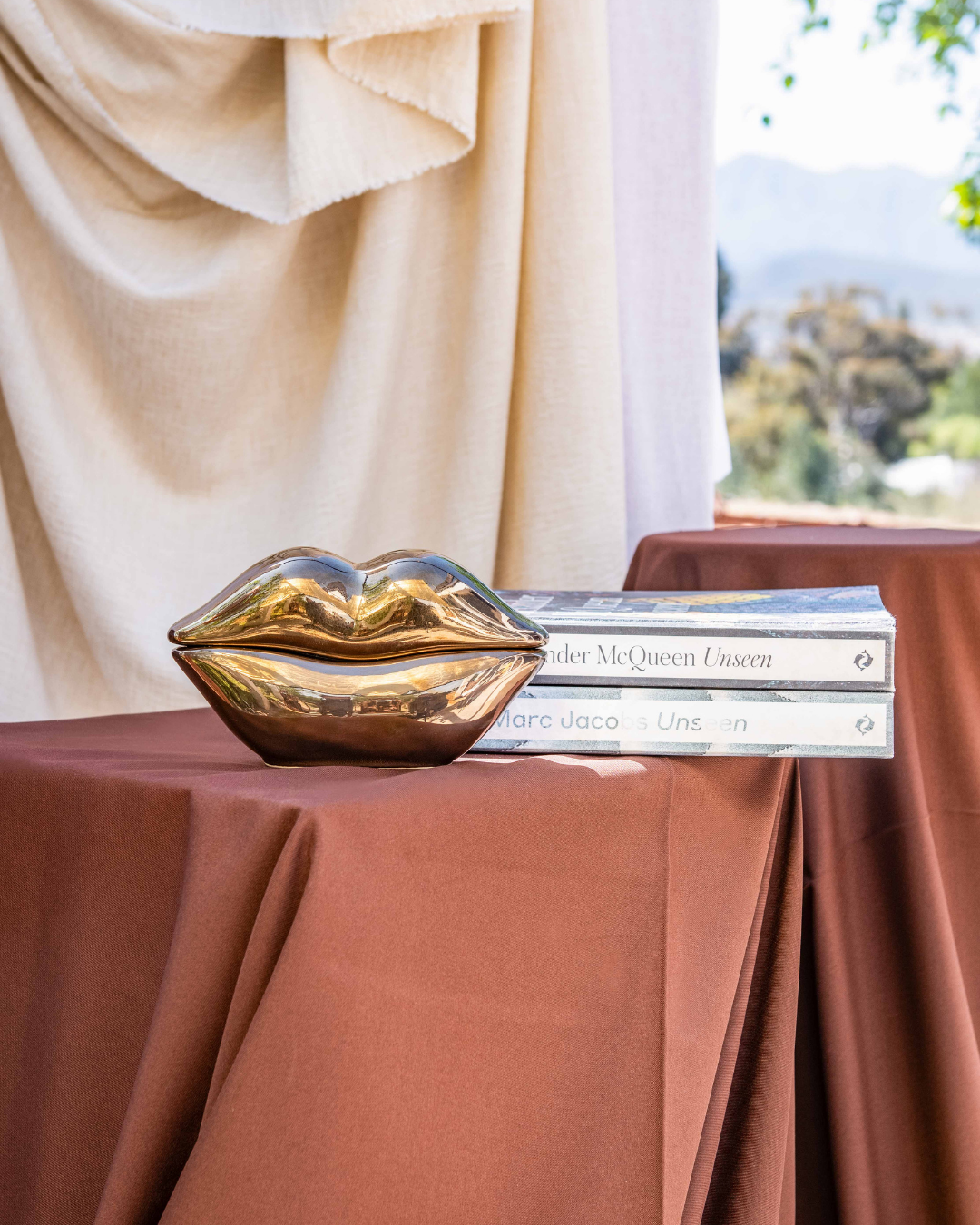 Gold lip-shaped object on a table with books underneath, against a beige curtain and window backdrop.