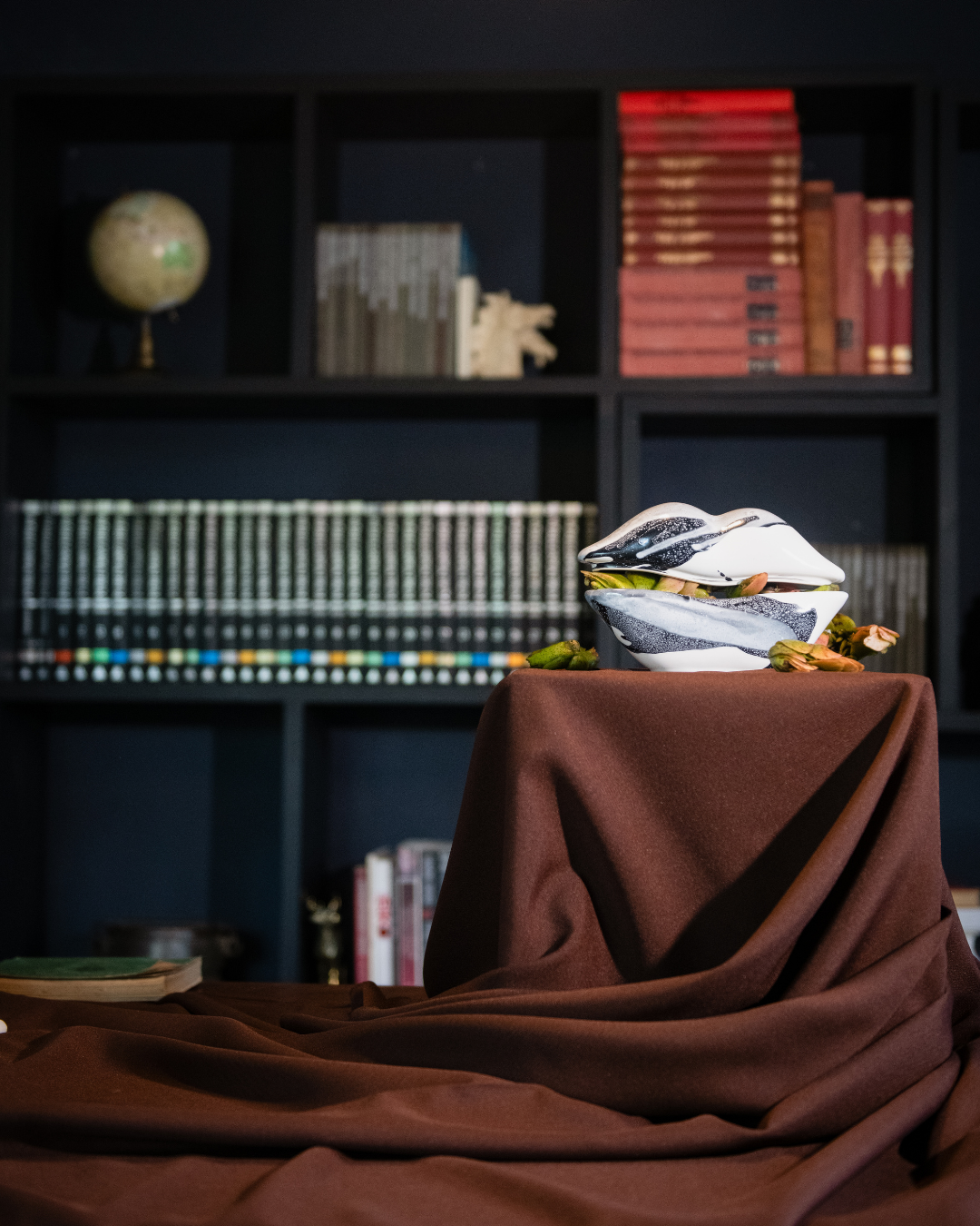 Decorative box on a brown cloth with a ceramic vessel in the shape of lips with a bookshelf in the background