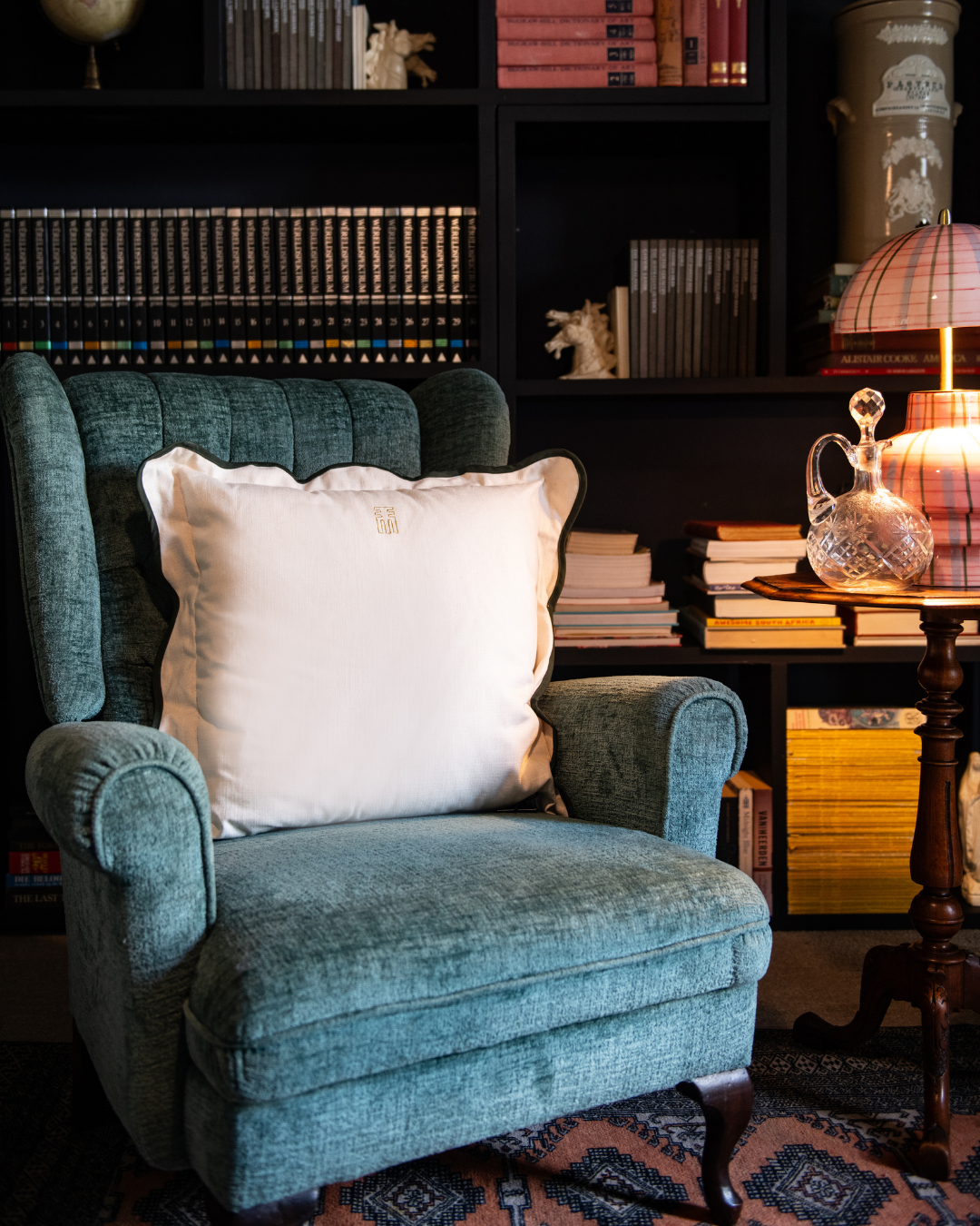 Blue armchair with a white Scallop pillow in a room with books and a lamp.