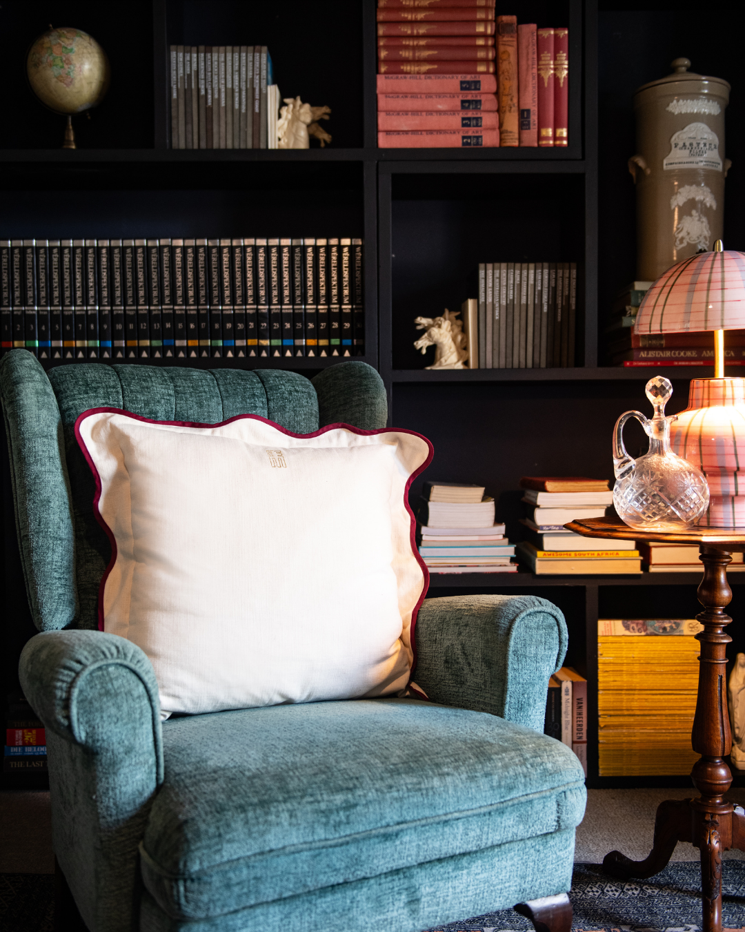 Blue armchair with a white Scallop pillow in a room with bookshelves and a lamp.