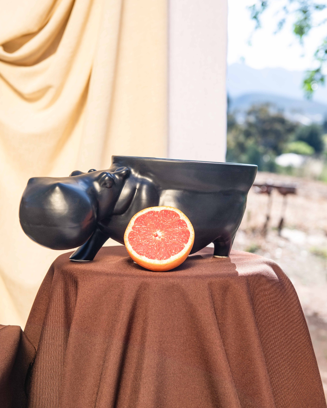 Decorative setup with ceramic hippo's with bowls attached to them turning them into fruit bowls on brown tablecloths, a white vase, and fruit bowl against a neutral curtain.