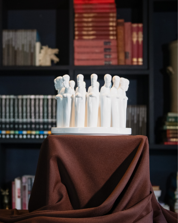 White sculptured praying men in a circle placed on a bowl on a brown cloth with a dark bookshelf background
