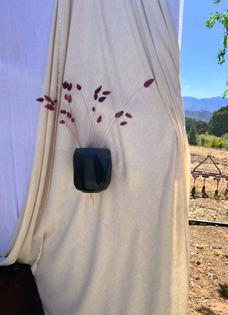 Black geometric cermaic vase attached to a white curtain with dried plants, outdoors.