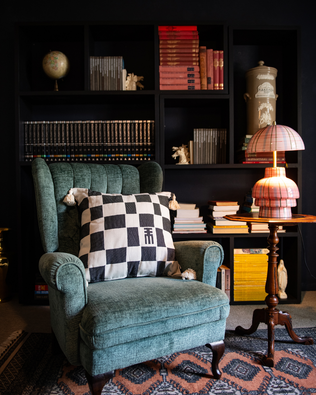 Green armchair with a checkered pillow in a room with bookshelves and a lamp.