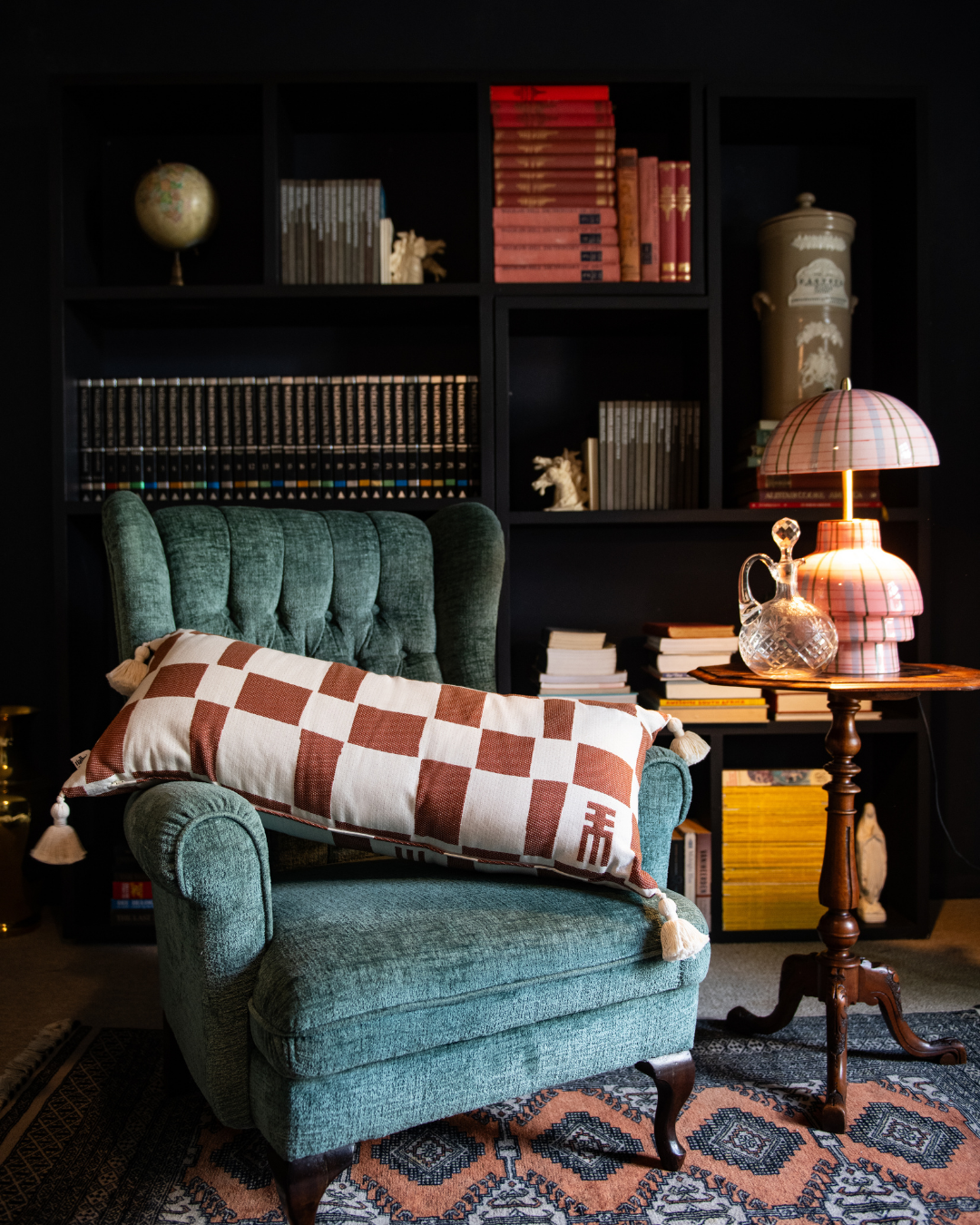 Green armchair with a checkered pillow in a room with a bookshelf and table lamp.