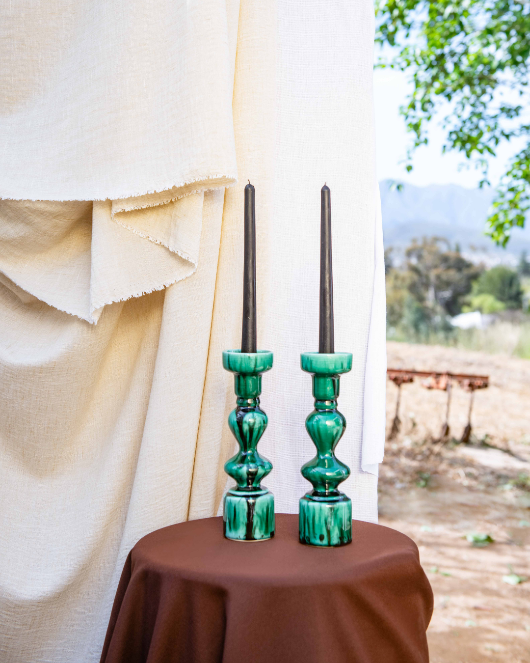Two green candle holders with black candles on a brown tablecloth against a white curtain and outdoor background.