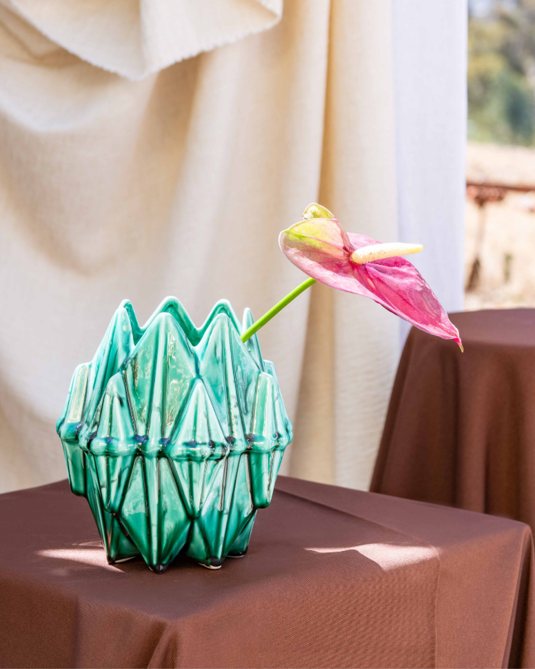 Green textured vase on a brown tablecloth with a pink flower against a beige curtain backdrop