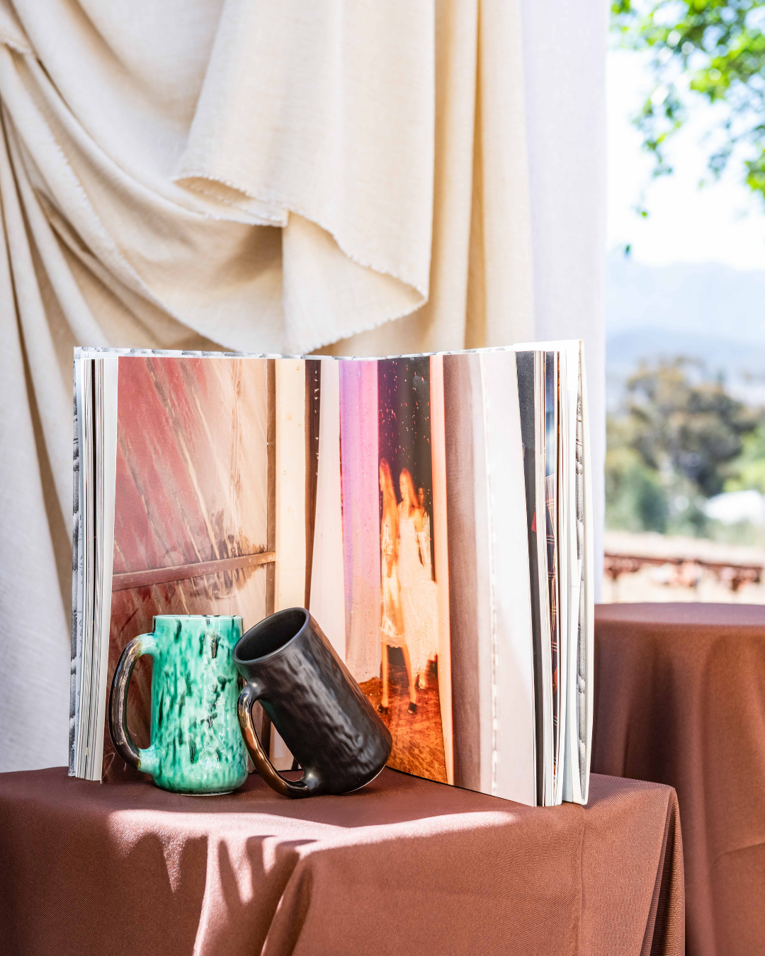 Open book with handmade ceramic mugs on a table with a curtain and outdoor view in the background