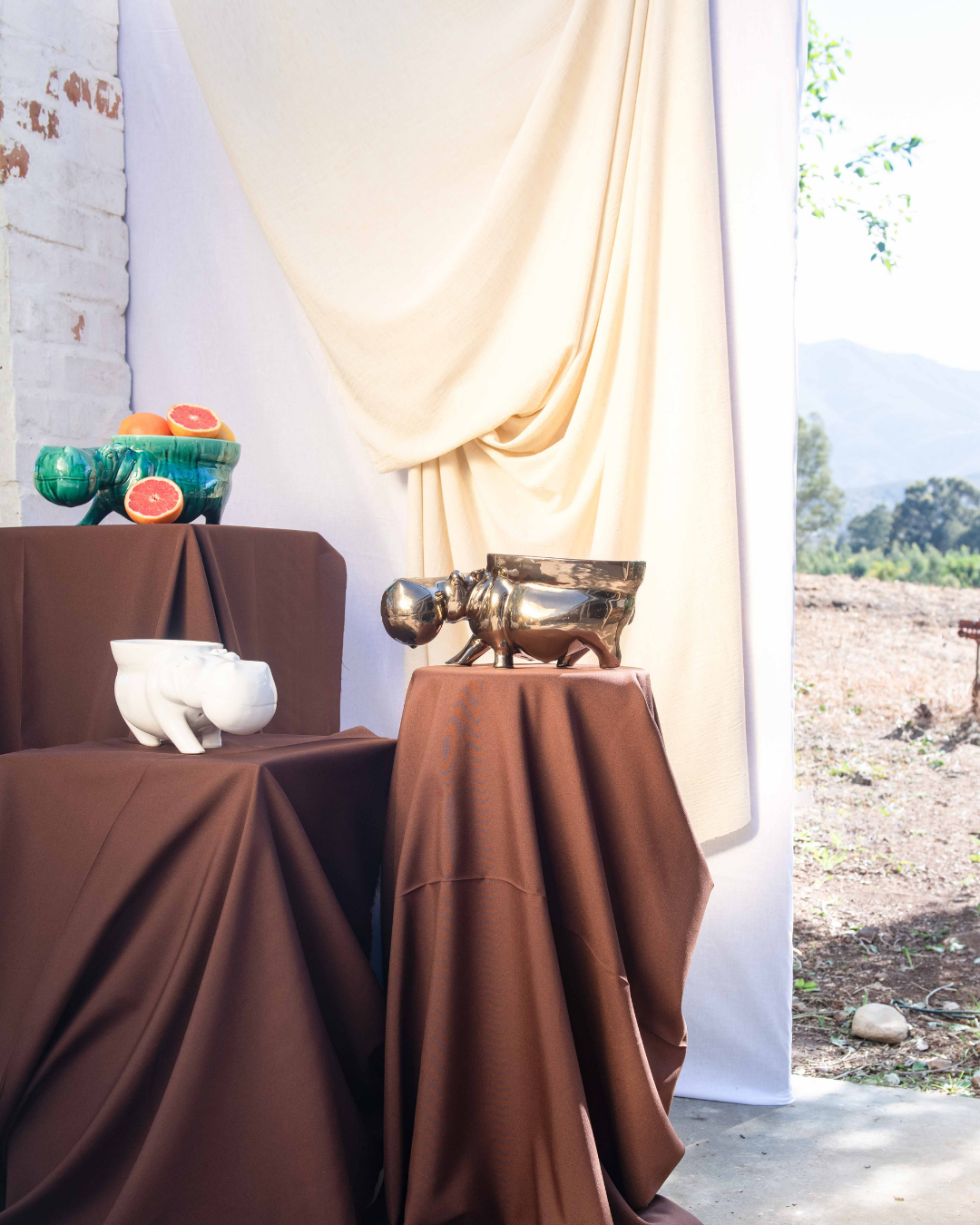 Decorative setup with ceramic hippo's with bowls attached to them turning them into fruit bowls on brown tablecloths, a white vase, and fruit bowl against a neutral curtain.