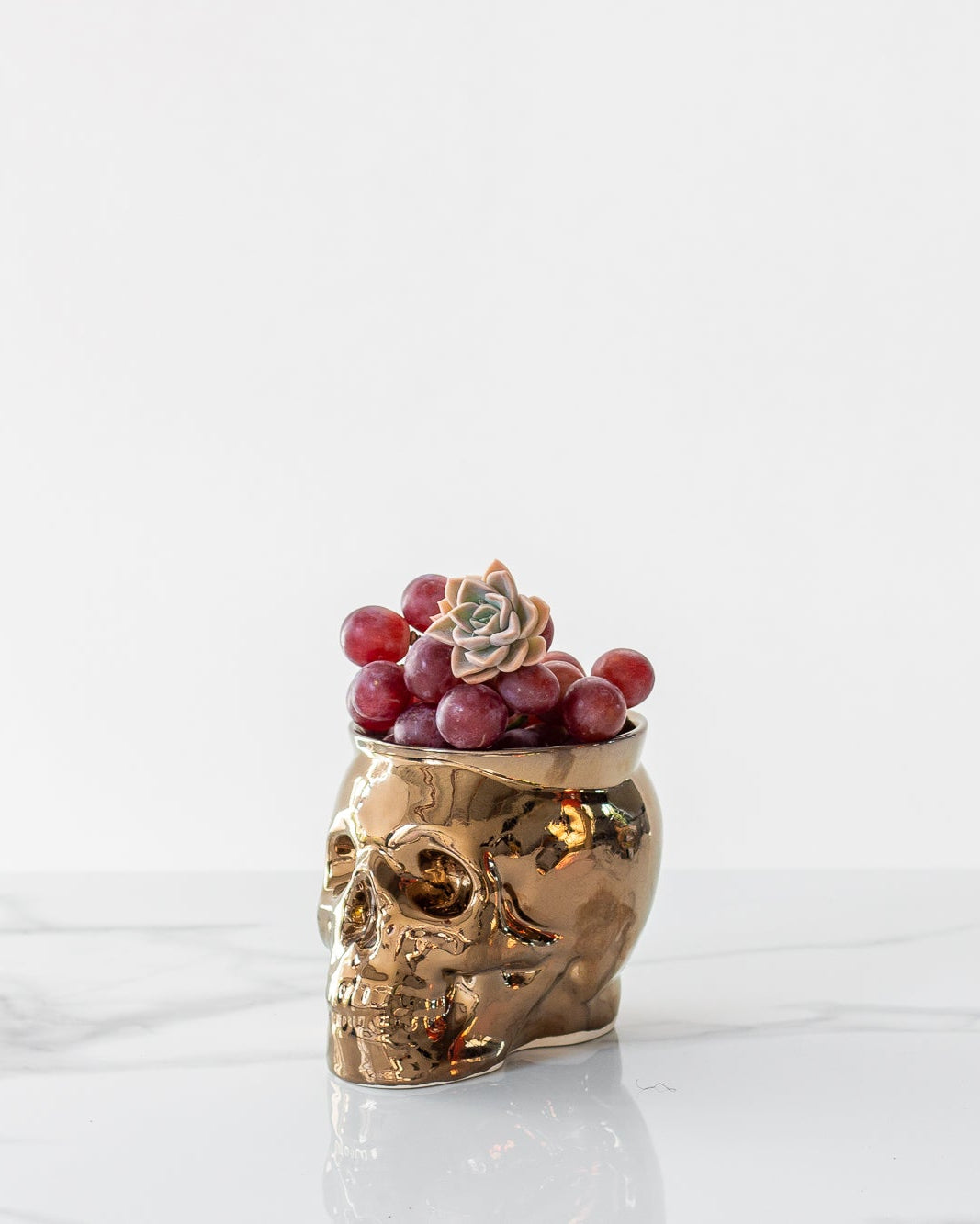 Gold skull-shaped container with grapes on a white marble surface