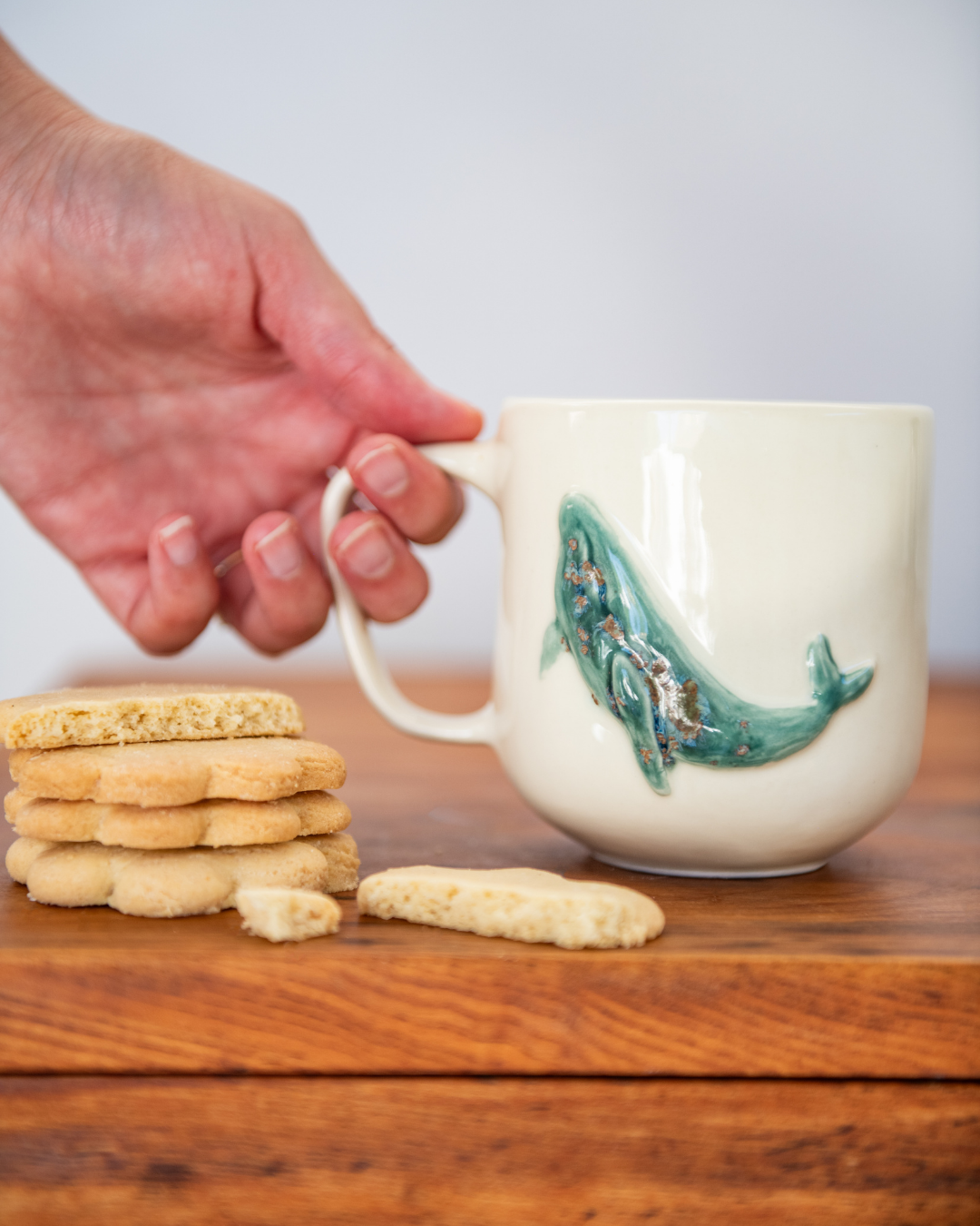 Hand holding a mug with a fish design next to a stack of cookies on a wooden surface.