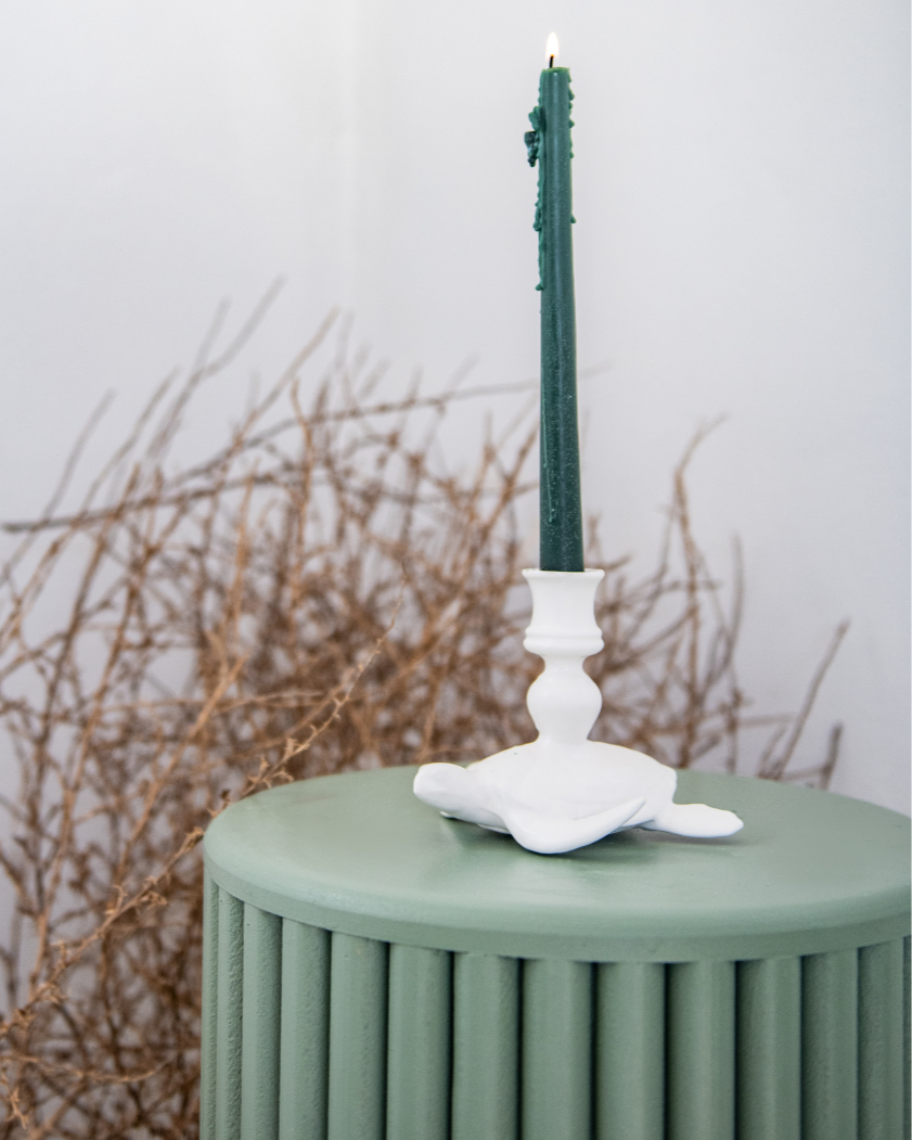 Decorative matt white sea turtle candle stick holders including a green candlestick, on an orange cushion against a white wall. 