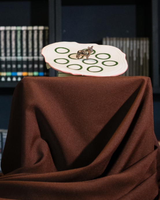 Dark room with a bookshelf and draped fabric covering and showing off a vintage shaped ceramic plate that is being hand painted by rialheim with a French Bull dog in Bronze in the middle of the plate