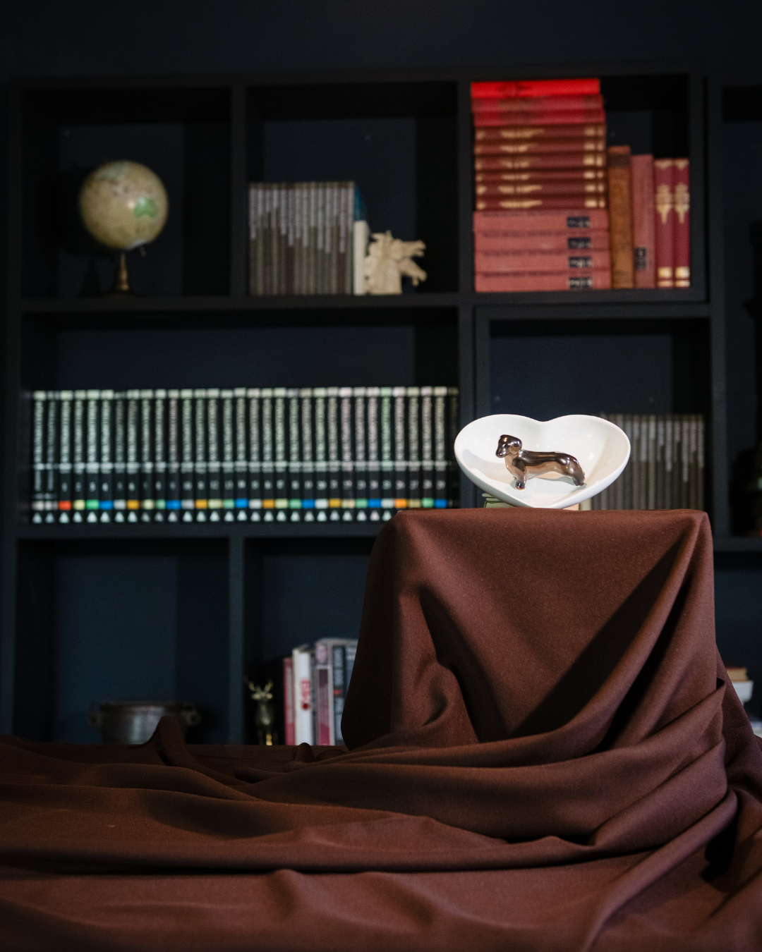 A gloss white bowl in the shape of a heart with one bronze Dashund glazed to the surface and a brown cloth draped over a table in the foreground.