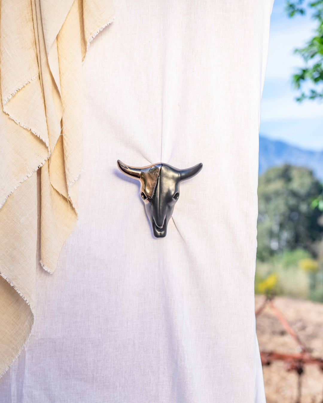 Metallic bull head pendant on a white fabric background with blurred outdoor scenery.