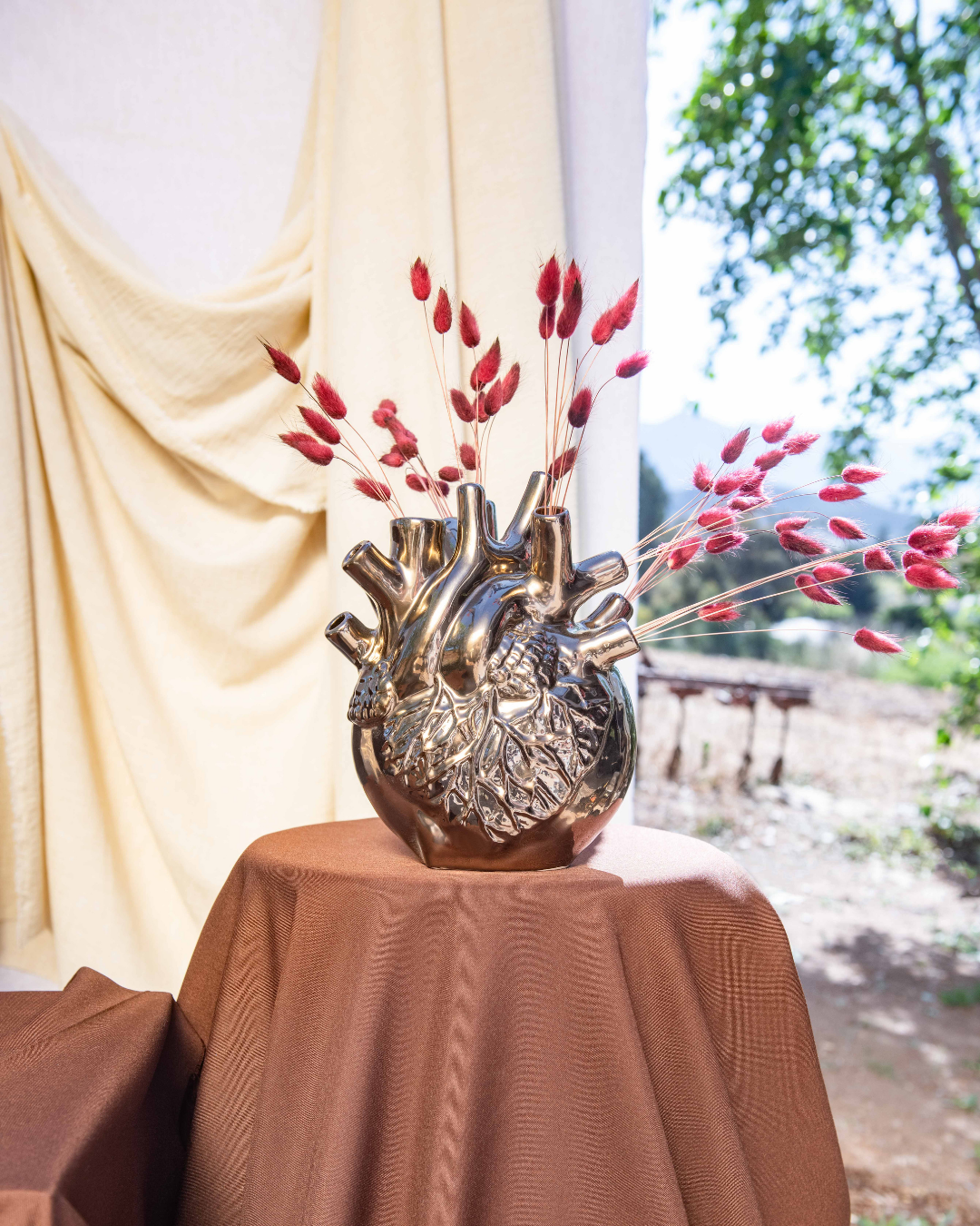Decorative bronze vase shaped like a heart with red branches on a table outdoors.
