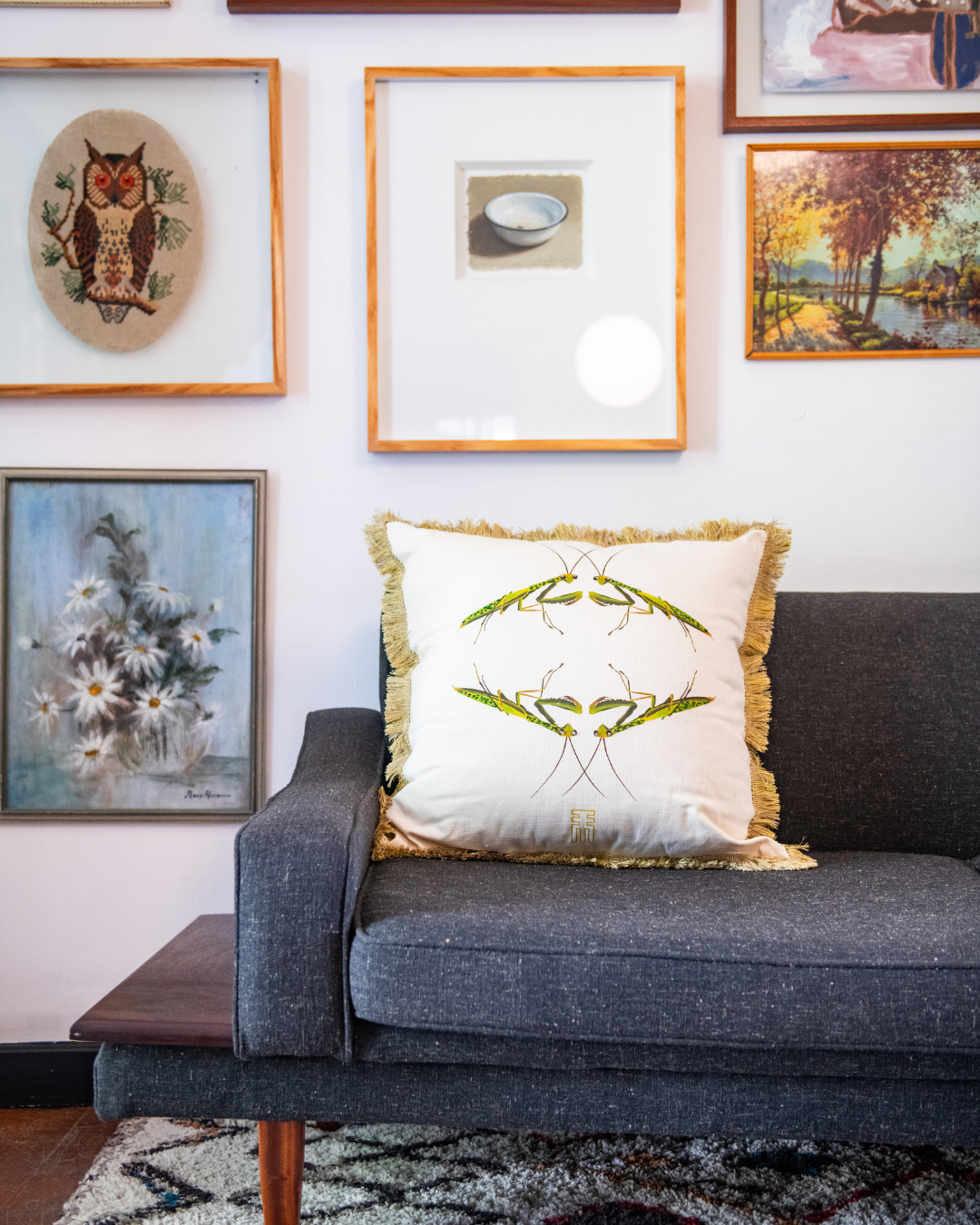 Decorative pillow on a gray sofa with framed artwork on the wall. there are 2 x pray mantises on the scatter cushion
