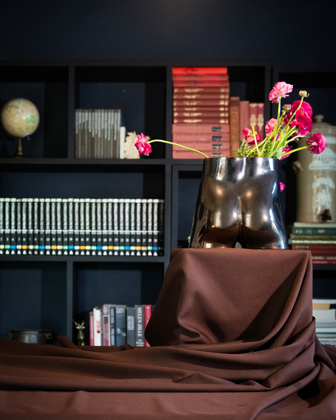 Decorative setup with a metallic bronze bum vase and flowers in front of a bookshelf.