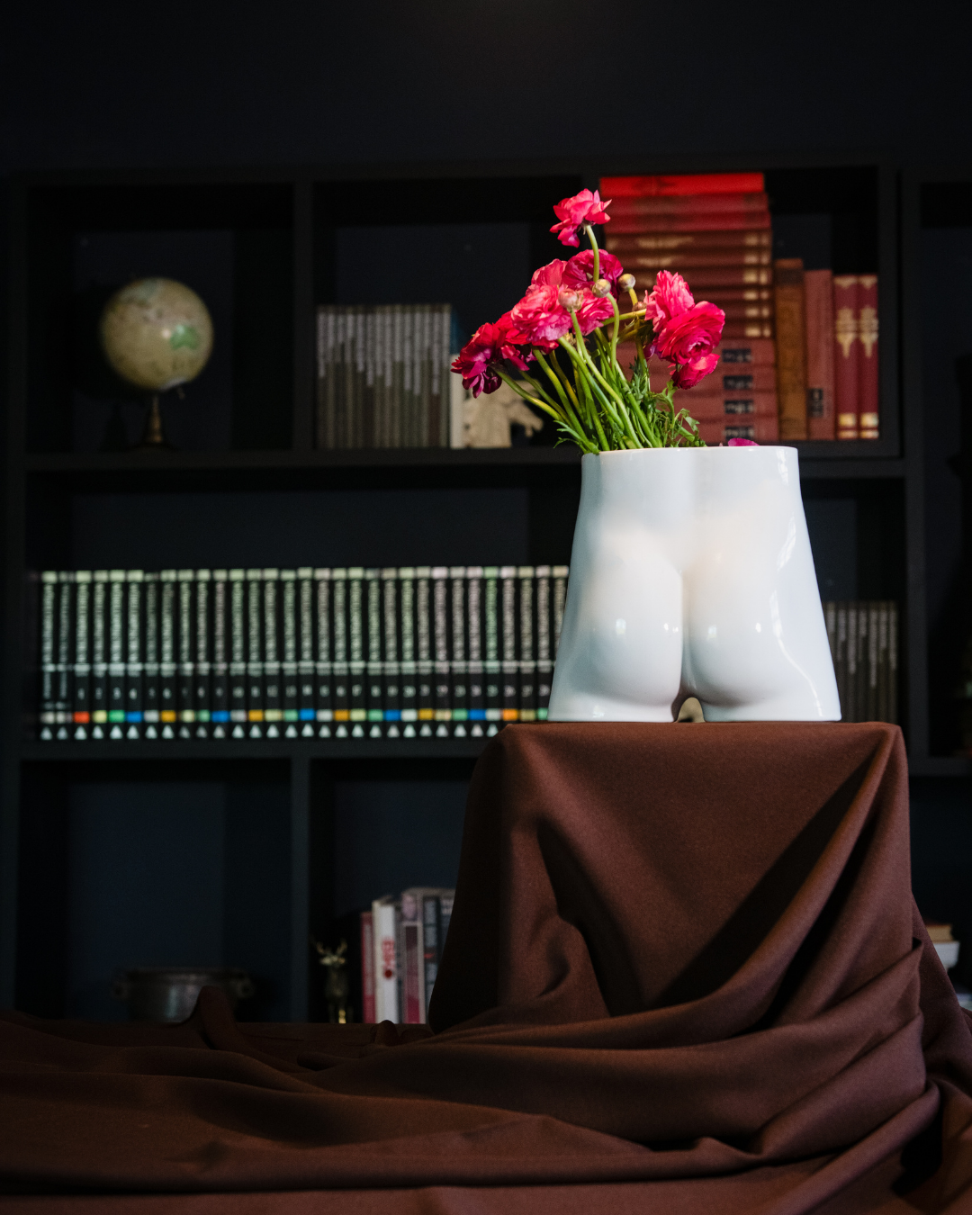 White vase shaped like a female or male bum with pink flowers against a dark bookshelf background