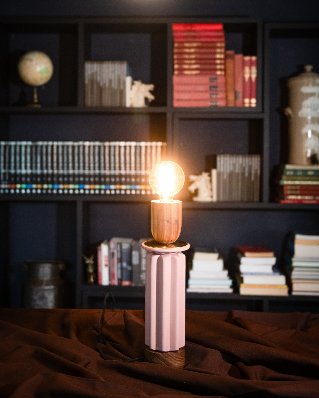 Table lamp with a lit bulb on a dark surface in front of a bookshelf.
