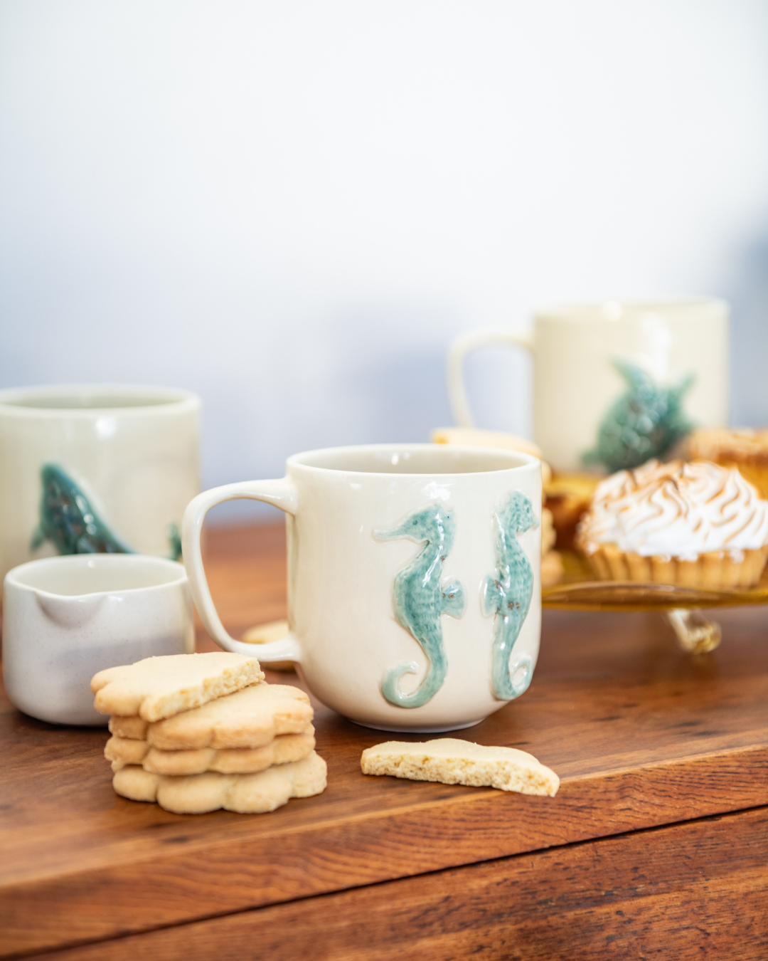 Mugs with seahorse designs on a wooden surface with cookies and a cake.