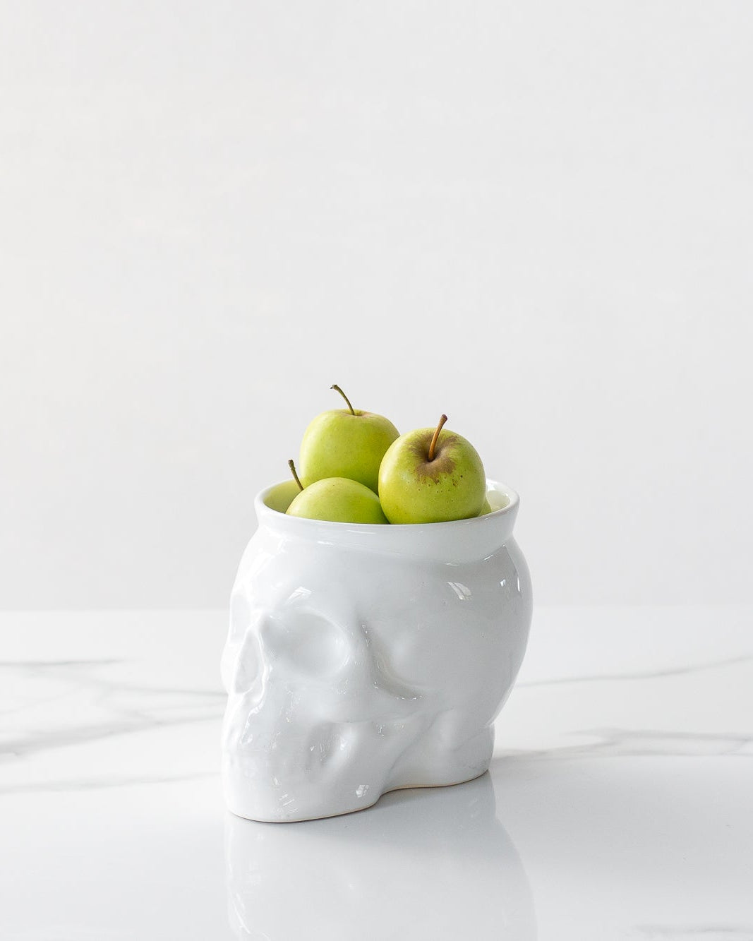 White ceramic skull-shaped vase with green apples on a white marble surface