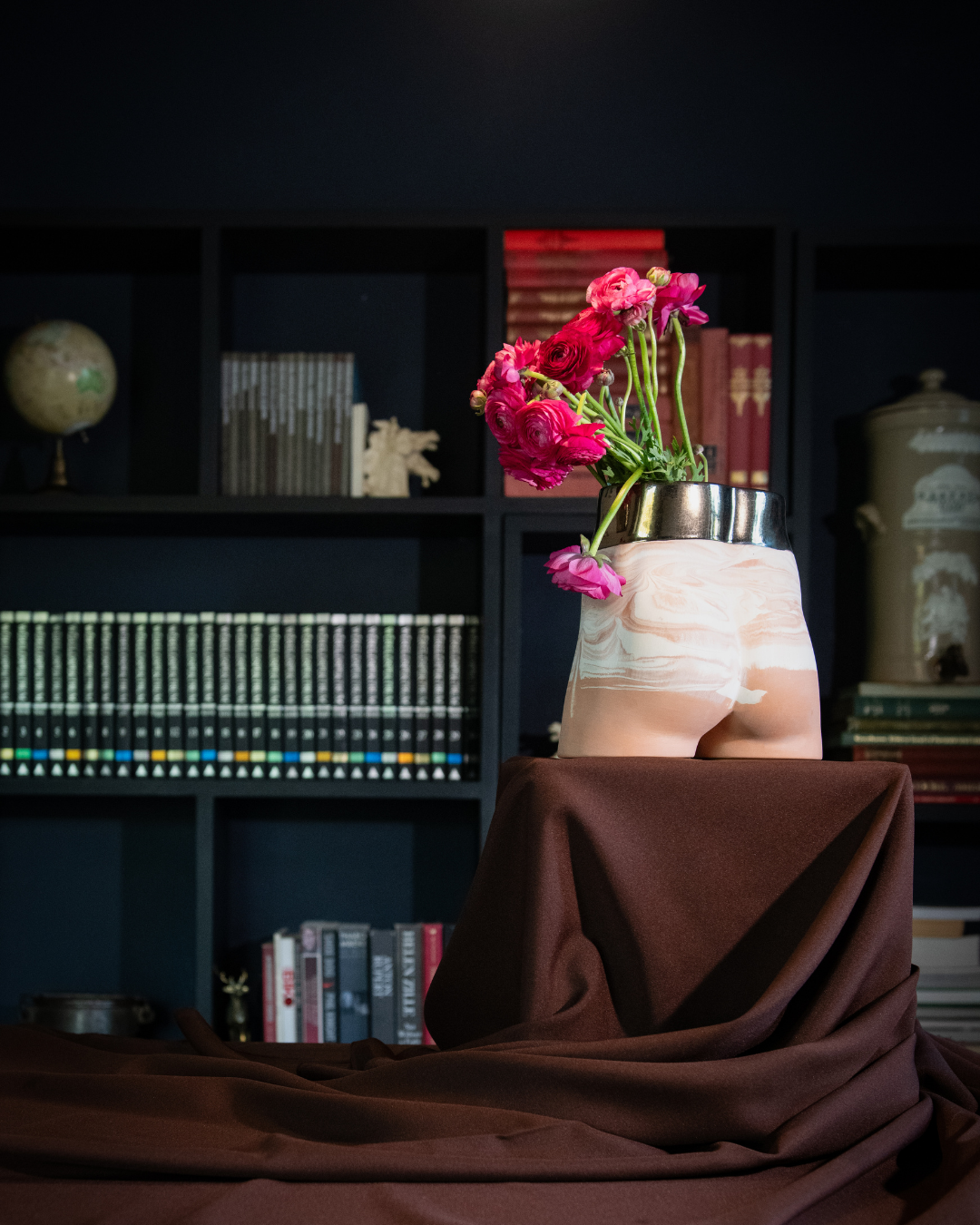 Decorative bum vase with pink flowers on a tablecloth in front of a bookshelf.