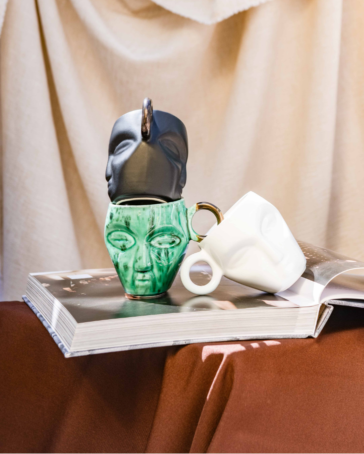 three rialheim mugs with ethic faces carved out on an open book with a beige curtain and outdoor view in the background.