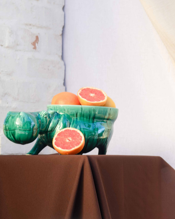 Decorative setup with ceramic hippo's with bowls attached to them turning them into fruit bowls on brown tablecloths, a white vase, and fruit bowl against a neutral curtain.