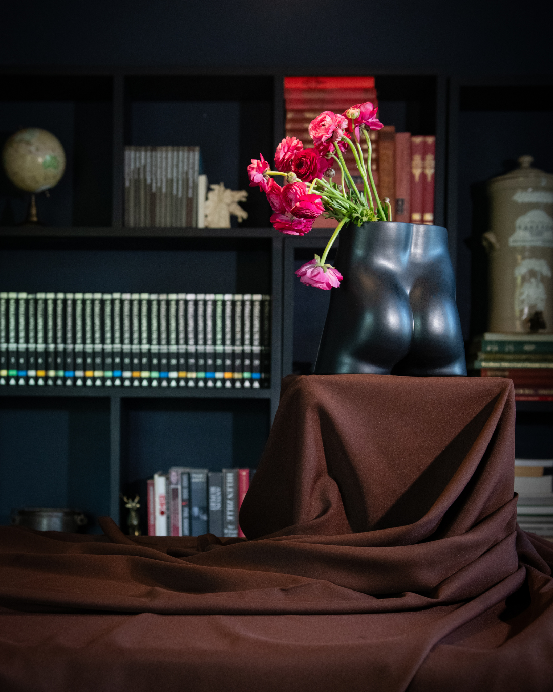 Rialheim Bum Vase in matt black with pink flowers on a draped brown cloth in front of a bookshelf.